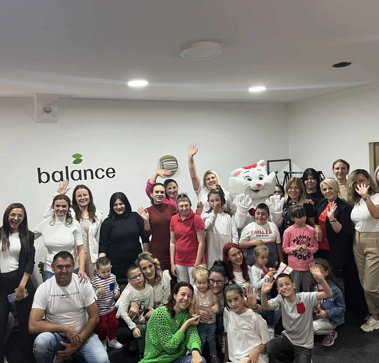 Grupa dece, roditelja i volontera Fondacije Balans sa maskotom belog mačeta ispred zida sa logom balance; svi mašu i osmehuju se u zajedničkoj prostoriji.